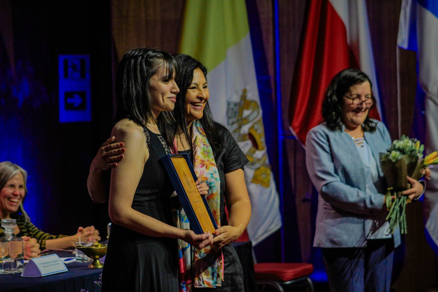 Mujeres en un acto de reconocimiento con bandera de fondo, una sostiene un reconocimiento y otra un ramo de flores.
