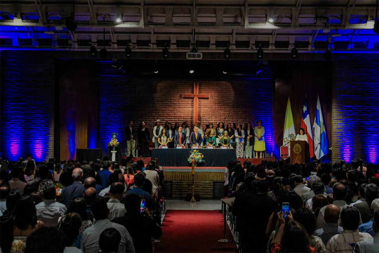 Escuela en una ceremonia religiosa con cruz y banderas, lleno de asistentes en un auditorio.
